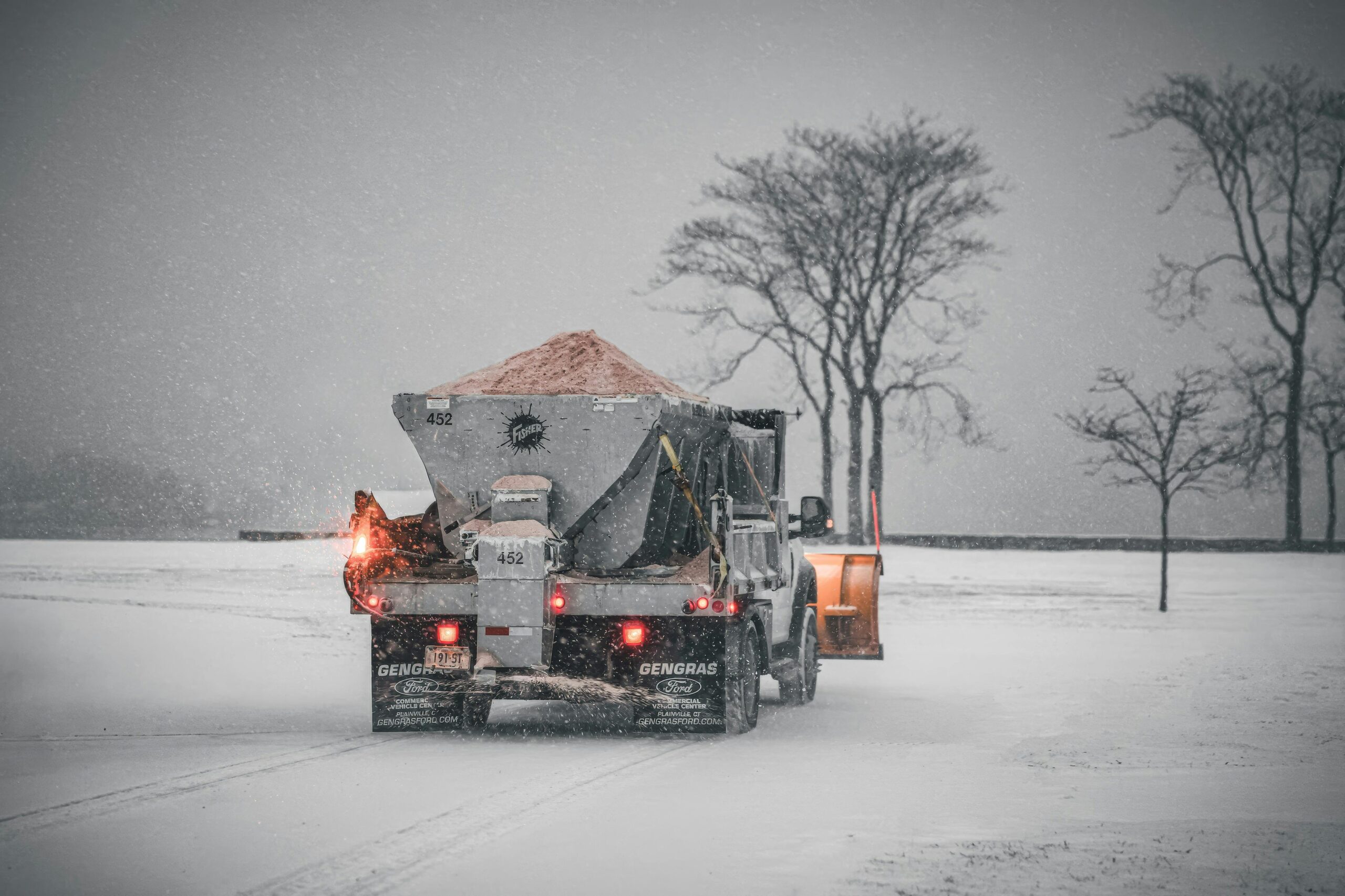 A snowplow truck spreading salt on a snowy road during a winter storm, with bare trees in the background.